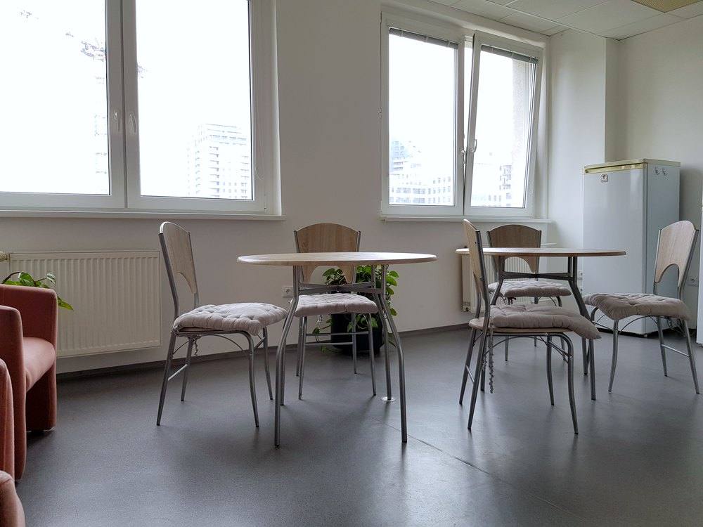Jedálenský kút s dvomi okrúhlymi stolmi, stoličkami, kreslom a chladničkou, okná s výhľadom na okolité budovy